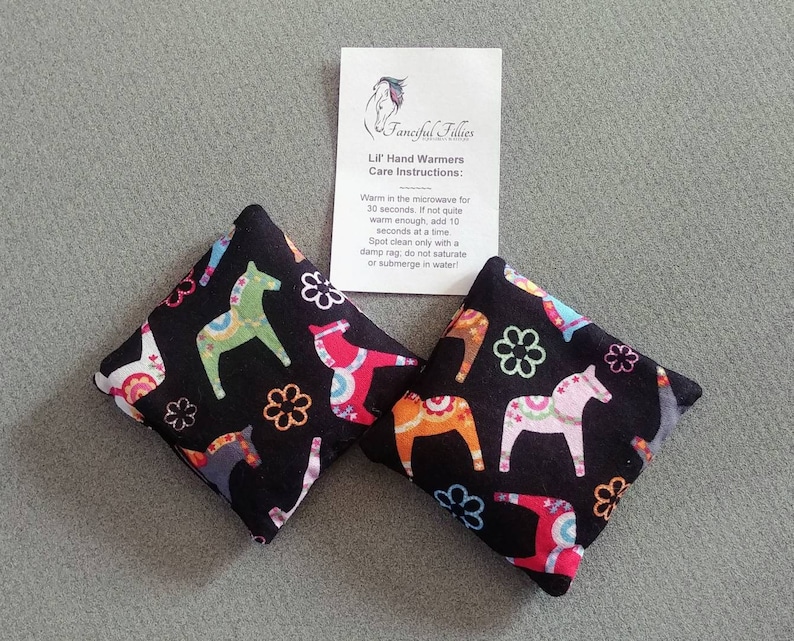 Puede incluir: Dos calientamanos de tela negra con un estampado de caballo escandinavo colorido. Los calientamanos est&aacute;n sobre una superficie gris con una tarjeta blanca que dice "Fanciful Fillies Lil' Hand Warmers Care Instructions: Warm in the microwave for 30 seconds. If not quite warm enough, add 10 seconds at a time Spot clean only with a damp rag, do not saturate or submerge in water!"