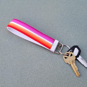 May include: A white key chain with a pink, orange, and yellow striped ribbon attached to a silver key ring with two keys. One key is gold and the other is black.