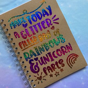 May include: A brown spiral notebook with a rainbow glitter design. The text on the notebook says "Make today a glitter filled day of rainbows & unicorn farts".