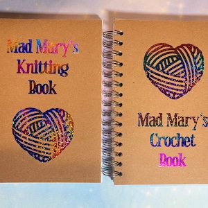 May include: Two brown spiral bound notebooks with rainbow glitter lettering and yarn heart designs. The left notebook says "Mad Mary's Knitting Book" and the right notebook says "Mad Mary's Crochet Book".