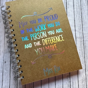 May include: A brown spiral notebook with a rainbow iridescent foil design that says "May you be proud of the work you do, the person you are, and the difference you make." The notebook also has the text "Miss Rx" in rainbow iridescent foil.