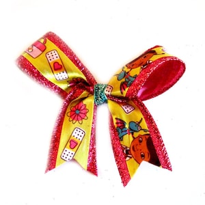 May include: A pink and yellow bow with a glitter finish. The bow features a cartoon character design with a bandage theme.