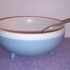 May include: A light blue and white bowl with three small legs, a brown rim, and a silver spoon inside. The bowl is sitting on a purple surface.