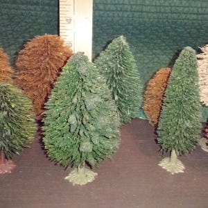 May include: A collection of miniature bottle brush trees in various colors, including green, brown, and tan. The trees have a textured, bristled appearance and are arranged on a dark surface. Some trees are cone-shaped, while others are rounded.