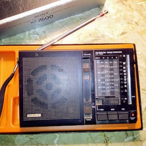 May include: A vintage black Sony ICF-7600 radio receiver with an extendable antenna, set in an orange tray. The radio features a tuning dial, volume control, and various buttons. The Sony logo is visible on the front.