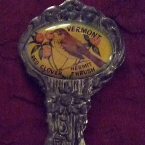May include: A silver-colored spoon with a decorative handle and an oval emblem. The emblem features a yellow background with a brown bird illustration, the words "VERMONT", "RED CLOVER", and "HERMIT THRUSH".