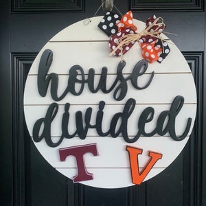 May include: A white round wooden sign with the text "house divided" in black script. The sign has two large letters, a maroon "T" and an orange "V" on the bottom. A black and white polka dot bow with orange and brown ribbon is at the top of the sign.