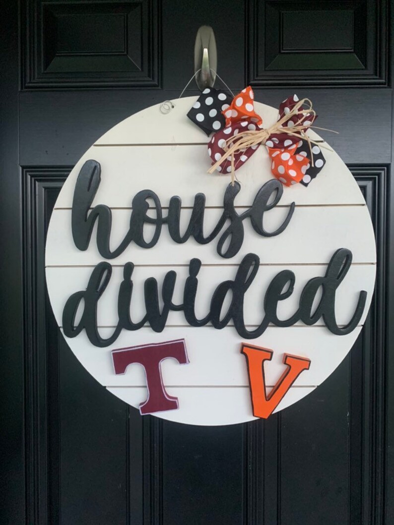 May include: A white round wooden sign with the text "house divided" in black script. The sign has two large letters, a maroon "T" and an orange "V" on the bottom. A black and white polka dot bow with orange and brown ribbon is at the top of the sign.