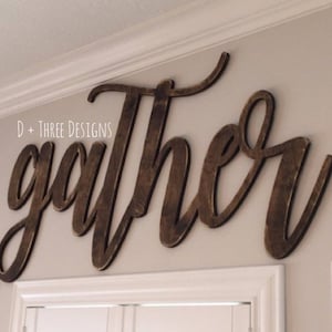 May include: A wooden sign with the word "gather" in a cursive font. The sign is painted in a dark brown color and is mounted on a white wall.