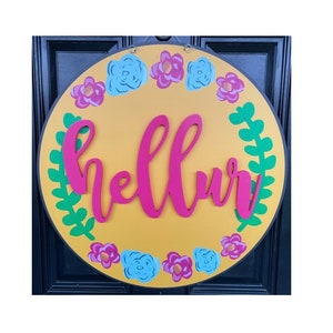 May include: A round, yellow wooden sign with a pink "hello" written in cursive. The sign is decorated with green leaves and pink and blue flowers.