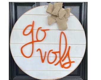 May include: A white wooden round door hanger with the words "go vols" in orange lettering. A burlap bow is attached to the top of the hanger.