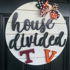 May include: A white round wooden sign with the text "house divided" in black script. The sign has a red and orange bow with white polka dots and the letters "T" and "V" in maroon and orange.