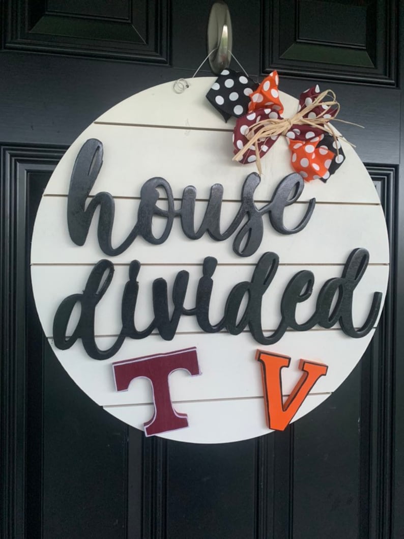 May include: A white round wooden sign with the text "house divided" in black script. The sign has a red and orange bow with white polka dots and the letters "T" and "V" in maroon and orange.