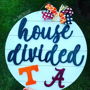 May include: A white round wooden sign with the text "house divided" in black script. The sign has a red and white polka dot bow and an orange and white patterned bow. The letters "T" and "A" are in orange and maroon, respectively.