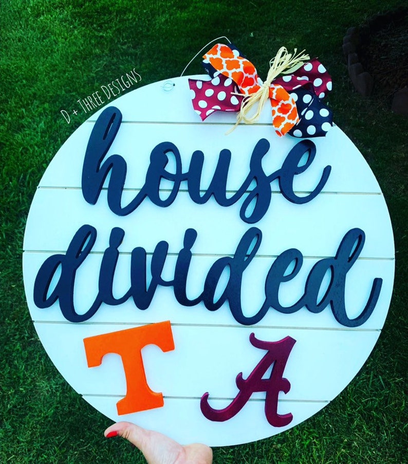 May include: A white round wooden sign with the text "house divided" in black script. The sign has a red and white polka dot bow and an orange and white patterned bow. The letters "T" and "A" are in orange and maroon, respectively.