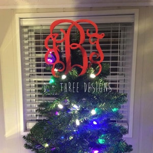 May include: A red wooden monogram Christmas tree topper with the letters "JBS" in a script font. The topper is on top of a green Christmas tree with white and multi-colored lights.