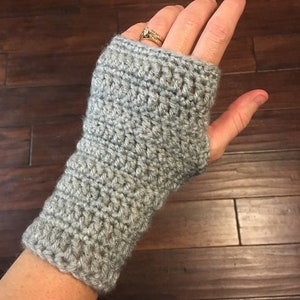 May include: A gray crocheted fingerless glove. The glove is made of a soft, textured yarn and covers the wrist and the base of the fingers, leaving the fingertips exposed. The crochet stitches are visible, creating a detailed pattern.