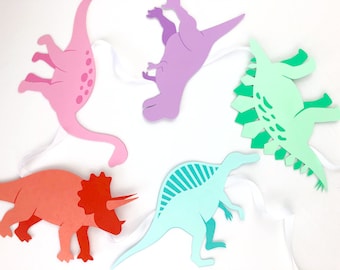 May include: A garland of five paper dinosaurs in different colors, including pink, purple, green, blue, and orange. The dinosaurs are cut out and strung together with white ribbon.