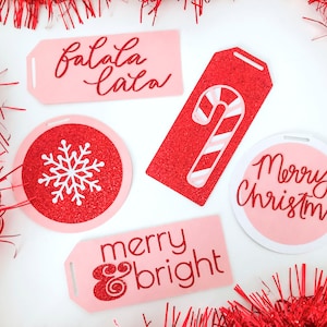 Puede incluir: Cinco etiquetas de regalo de Navidad de color rosa y rojo con diferentes diseños. Las etiquetas presentan el texto "Feliz Navidad", "Feliz y brillante", "Falala Lala" y un copo de nieve. Una etiqueta presenta un bastón de caramelo rojo y blanco.