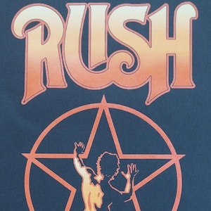 May include: A dark blue t-shirt featuring the word RUSH in a gradient orange font above a graphic of a person standing inside a star. The design includes the text 2026 SRO/YYZ RUSH Under License to Merch Traffic printed at the bottom of the graphic.