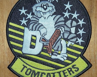 May include: A black and yellow embroidered patch with a cartoon raccoon holding a gun. The raccoon is wearing a striped shirt and has a big smile. The patch says "Tomcatters" in yellow letters.