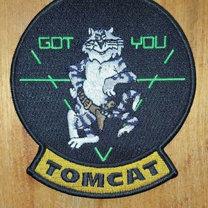 Puede incluir: Un parche bordado negro y verde con un gato blanco que lleva un cinturón y sostiene una pistola. El gato está en un triángulo con las palabras "GOT YOU" arriba y "TOMCAT" abajo.