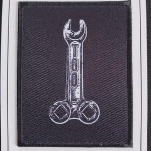 May include: A rectangular black fabric patch featuring a white distressed graphic of a wrench with the word TOOL printed vertically along the handle. The patch is mounted on a white rectangular display card with a top hole for hanging.
