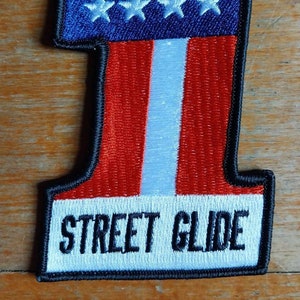 Puede incluir: Un parche bordado con forma de número uno, en rojo, blanco y azul. La parte superior del parche es un campo azul con estrellas blancas. La parte inferior del parche es de rayas rojas y blancas con una raya blanca en el centro. El texto "STREET GLIDE" está bordado en negro sobre un fondo blanco en la parte inferior del parche.