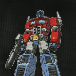 Könnte beinhalten: Gestickter Aufnäher von Optimus Prime, einem roten, blauen und silbernen Robotercharakter. Der Aufnäher hat detaillierte Nähte, eine schwarze Pistole und ist vor einem schwarzen Hintergrund platziert. Der Roboter steht.
