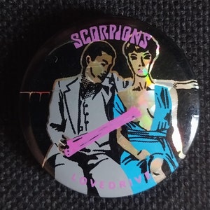 Op de afbeelding: Een zwart-witte knop met een holografische afwerking met een band genaamd "SCORPIONS" met de woorden "LOVE DRIVE" onderaan. De knop toont een man en een vrouw in een zwart-witte illustratie met een roze object in de hand van de vrouw.