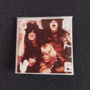 May include: A square pin featuring a group of four people with dark hair and makeup, with the word "CURE" in white letters. The image has a vintage, concert-like aesthetic, with a dark background.