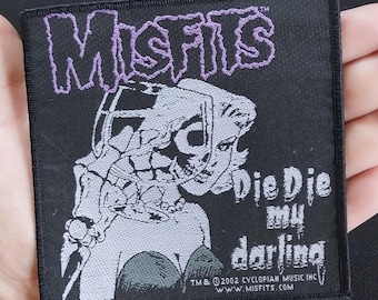 Puede incluir: Parche cuadrado negro con el nombre de la banda "Misfits" en morado. Un gráfico en blanco y negro de una mujer con rostro de calavera está debajo. El texto "Die Die my darling" también está presente. Hecho de tela.
