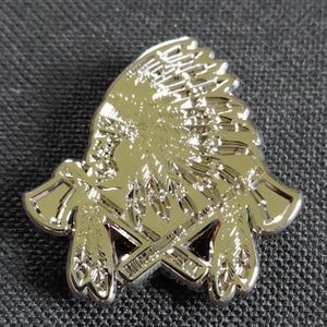 May include: A silver-colored metal pin featuring a Native American headdress and crossed tomahawks. The pin has detailed relief work and a shiny finish, with a classic design.