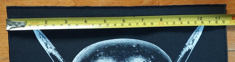 May include: Black rectangular patch with a white graphic design. A yellow tape measure shows the patch is approximately 10 inches long. The patch is on a wooden surface.