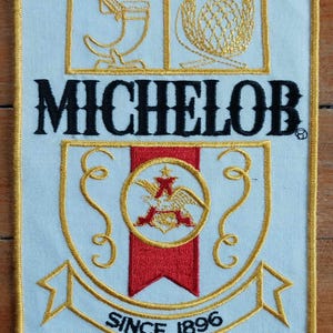 May include: Embroidered patch with the word "MICHELOB" in black, and gold and red accents. The patch features a shield design with the text "SINCE 1896". The patch has a light blue background and a gold border.