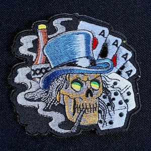 Peut inclure: Écusson brodé représentant un crâne portant un chapeau haut de forme bleu, fumant une cigarette, avec des cartes à jouer, des dés et une bouteille. Le crâne a des yeux verts. Sur fond noir.