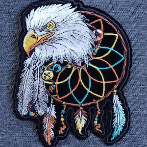 Può includere: Toppa ricamata raffigurante una testa d'aquila con piume bianche e becco giallo, integrata in un design acchiappasogni. L'acchiappasogni presenta accenti blu, arancioni e gialli, con piume pendenti. La toppa ha un bordo nero.