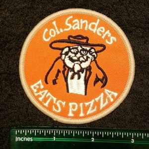 May include: Orange and white embroidered patch with the text "Col. Sanders Eats Pizza" and a cartoon image of Colonel Sanders wearing a hat and a white beard.