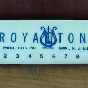 May include: A vintage off-white Royastone harmonica with blue lettering and a lyre logo. The text includes "PROLL TOYS INC." and "NWK., N. J. U.S.A." along with the numbers 1 through 10. The harmonica is on a wooden surface.