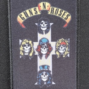 Pode incluir: Remendo retangular com o logotipo Guns N' Roses. O design inclui uma cruz branca com rostos de caveira e o nome da banda em uma faixa. O remendo tem uma borda e fundo pretos.