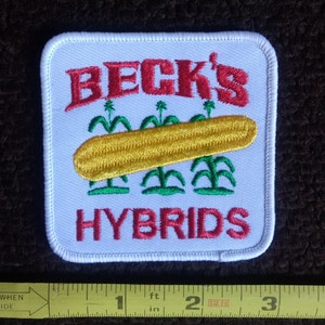 May include: White embroidered patch with red text that reads "BECK'S HYBRIDS". The patch features a yellow ear of corn with green corn stalks.