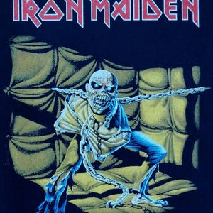 May include: Black t-shirt featuring the band name "Iron Maiden" in red block letters. The graphic depicts a skeletal figure with chains, blue eyes, and a yellow cloak, set against a yellow padded backdrop.