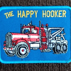 May include: A blue embroidered patch with the text "THE HAPPY HOOKER" in yellow lettering. The patch features a red, white, and silver tow truck.