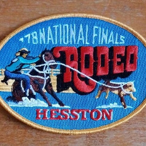 Può includere: Toppa ricamata con sfondo ovale blu e bordo dorato. La toppa presenta una scena di rodeo con un cowboy a cavallo, un cane e la parola "RODEO" in rosso. Il testo include "'78 NATIONAL FINALS" e "HESSTON".