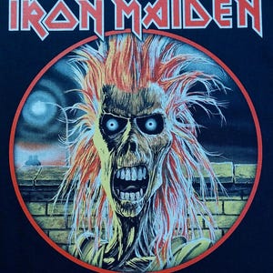 May include: Black t-shirt with the Iron Maiden logo in red and a circular graphic. The graphic depicts a zombie-like figure with orange hair and blue eyes, set against a night scene with a brick wall.