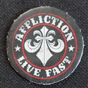 May include: A black and red circular patch with the word "AFFLICTION" in white lettering around the edge. A white fleur-de-lis is in the centre with the words "LIVE FAST" in white lettering around the bottom edge.