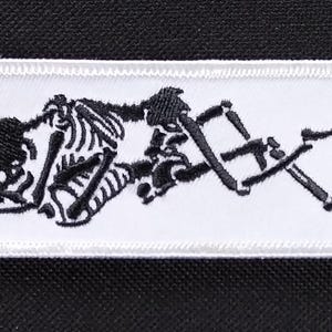May include: White embroidered patch with a black skeleton figure lying down. The skeleton is in a relaxed pose with its arms and legs crossed.