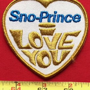 May include: A white heart-shaped patch with gold embroidered trim. The words "Sno-Prince" are embroidered in blue and the words "LOVE YOU" are embroidered in gold.