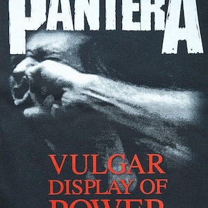 Peut inclure: T-shirt noir avec le nom du groupe "PANTERA" en lettres blanches en haut. En dessous, une image en niveaux de gris d'un poing serré. Les mots "VULGAR DISPLAY OF POWER" sont imprimés en rouge. Informations sur le copyright en bas.