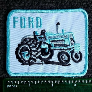 Op de afbeelding: Een blauwe en witte geborduurde patch met het woord "FORD" en een blauwe tractor met zwarte wielen.